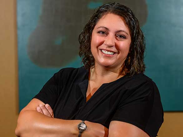 Lindsay Chichester - a woman with dark hair smiles and crosses her arms. She is wearing a dark shirt, a watch, and standing in front of a green background.